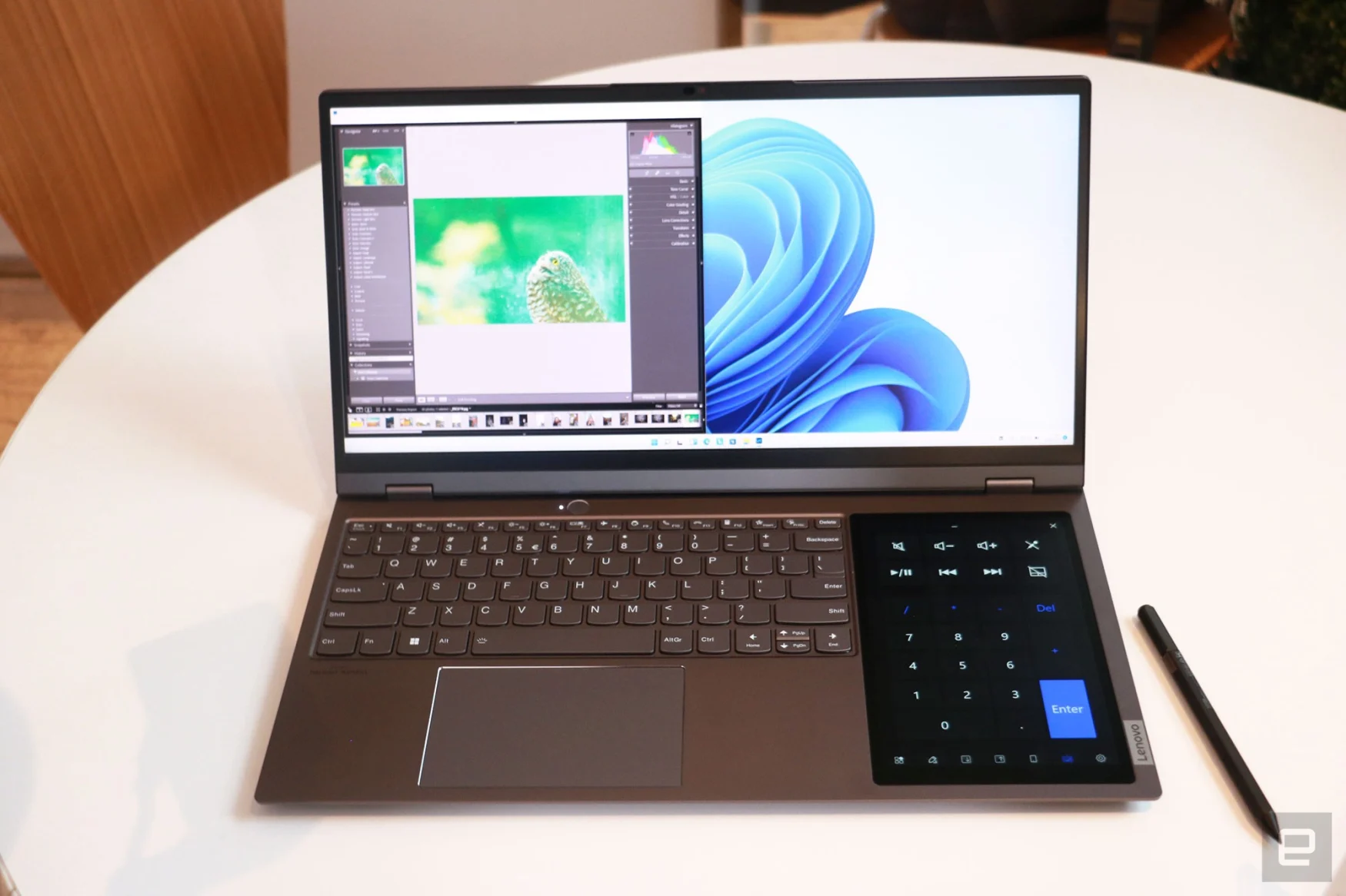 The Lenovo ThinkBook Plus Gen 3 sitting on a round white table with a stylus next to it. The laptop shows Adobe Lighthouse taking up the left half of the main display, while an oversized numpad appears on the 8-inch screen to the right of the keyboard.