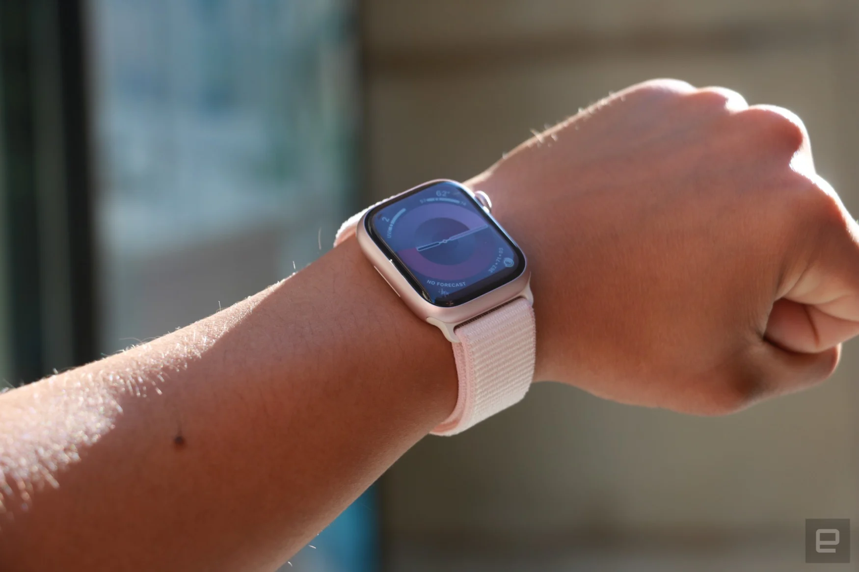 Apple Watch Series 9, havada bir bilek üzerinde, Digital Crown'dan yansıyan güneş ışığıyla birlikte ana ekranı gösterir.