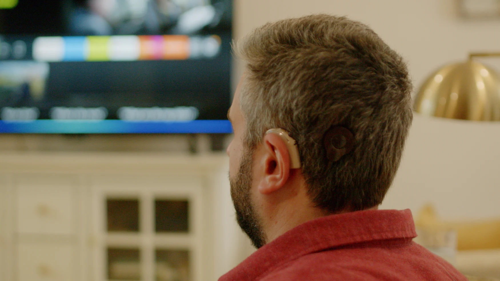 The back left of a bearded man’s head watching TV in his living room. He is wearing an assistive hearing device.