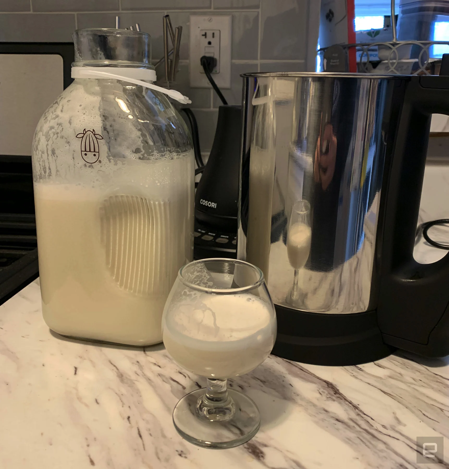 The Almond Cow plant milk maker next to a jug of homemade cashew milk and a glass with homemade cashew milk on a marbled countertop.