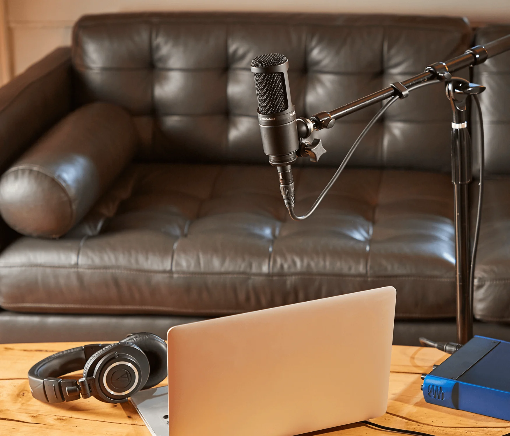 An Audio-Technica AT2020 condenser mic is on a stand above a coffee table with a laptop, with a leather couch in the background.