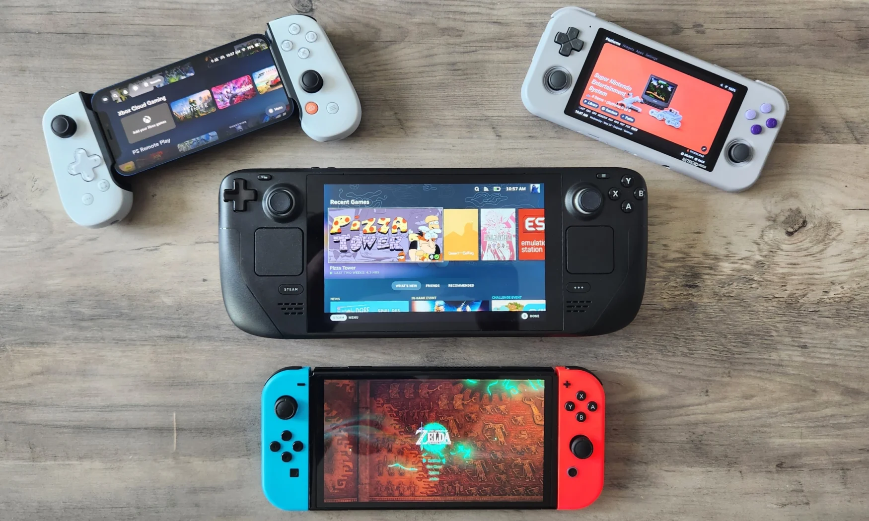 A collection of gaming handhelds rest on a wooden tabletop. The handhelds include the Nintendo Switch - OLED Model, Valve Steam Deck and the Retroid Pocket 3, as well as an iPhone 12 mini hooked up to a Backbone One mobile game controller.