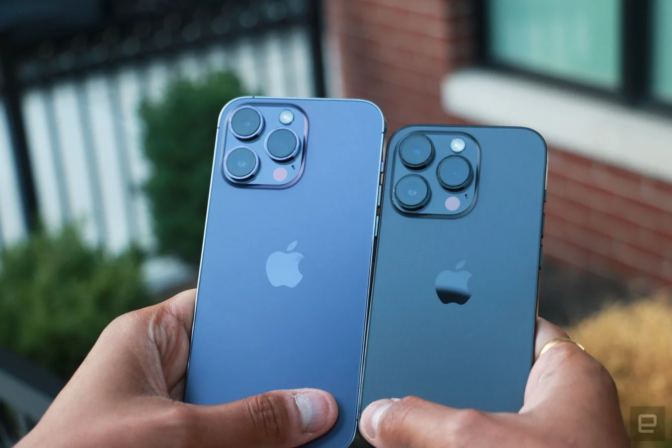Das iPhone 14 Pro Max und das iPhone 14 Pro werden in zwei Händen gehalten und mit nach außen gerichteten Rückkameras nebeneinander platziert.
