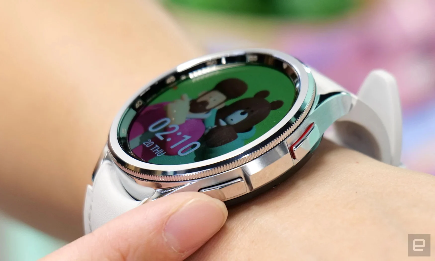 Closeup photo of a Samsung Galaxy Watch 6 smartwatch on a person's wrist. A finger is touching one of the hardware buttons as an image of a man with a teddy bear displays on its screen.