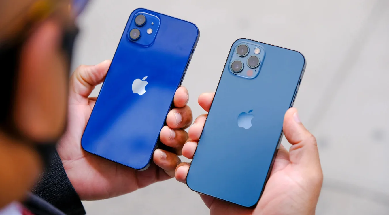 A person holding a blue iPhone 12 and blue iPhone 12 pro in each hand, with the devices' rear cameras facing up. 