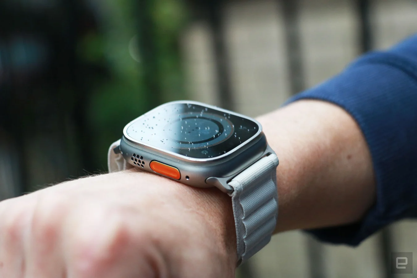 Off-Winkel-Ansicht der Apple Watch Ultra mit alpiner Schlaufe am Handgelenk einer Person.  Aus dieser Perspektive sind die orangefarbene Aktionstaste und das Lautsprechergitter am linken Rand sichtbar, während der Bildschirminhalt abgedunkelt ist.