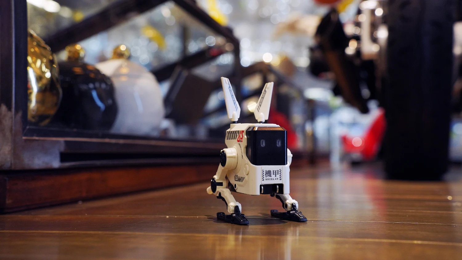 A cute robot charger (mech-inspired) sitting on a wooden desk. Various blurred objects are behind it.
