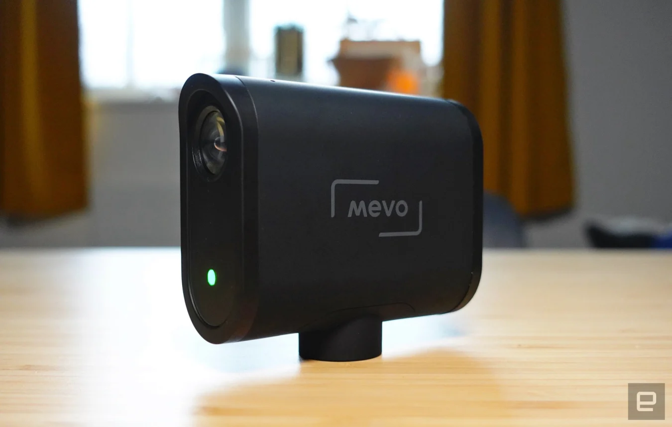The Logitech Mevo livestreaming camera sitting out on a wooden table with a window in the background.