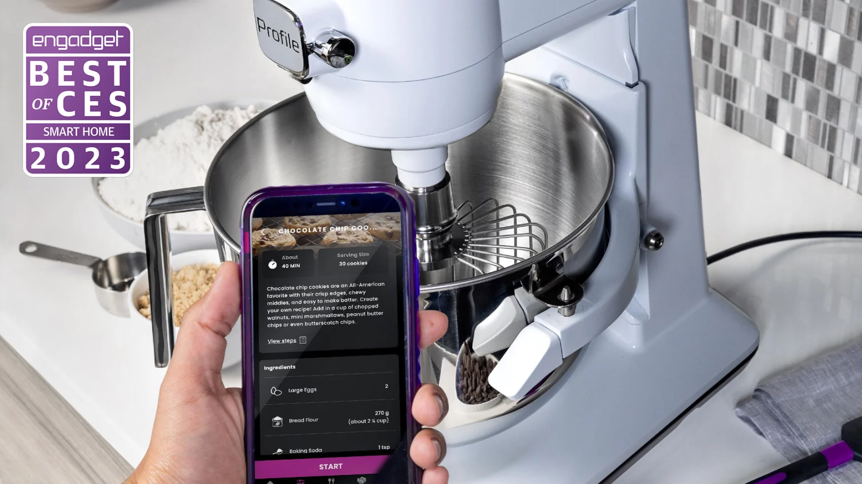 A cropped photo showing someone's hand holding a smartphone in front of a white GE Profile Smart Mixer on a white kitchen counter with grey tiled backsplash.