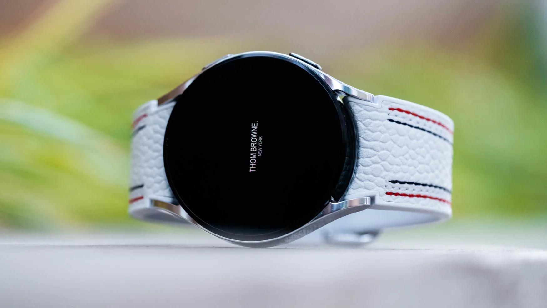 A Thom Browne edition of the Samsung Galaxy Watch 4 laying on its side with greenery in the background.